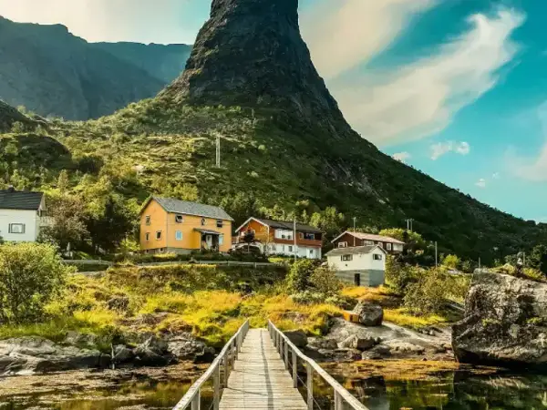 A scenic mountain village with colorful houses nestled beneath a steep peak, connected by a wooden footbridge over calm water