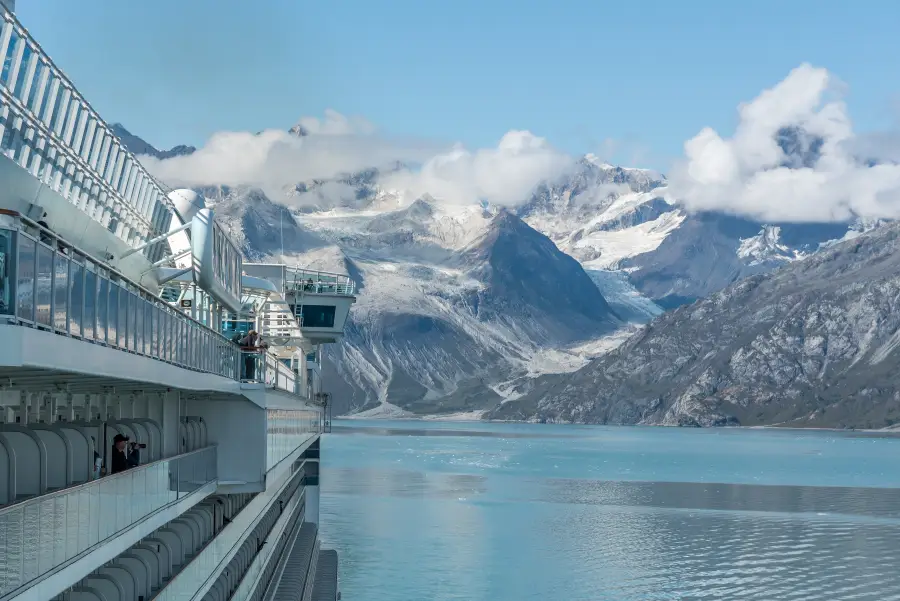 Alaska cruise ship balconies overlooking glacial Glacier Bay waters with snow-capped mountains and flowing glaciers beyond.