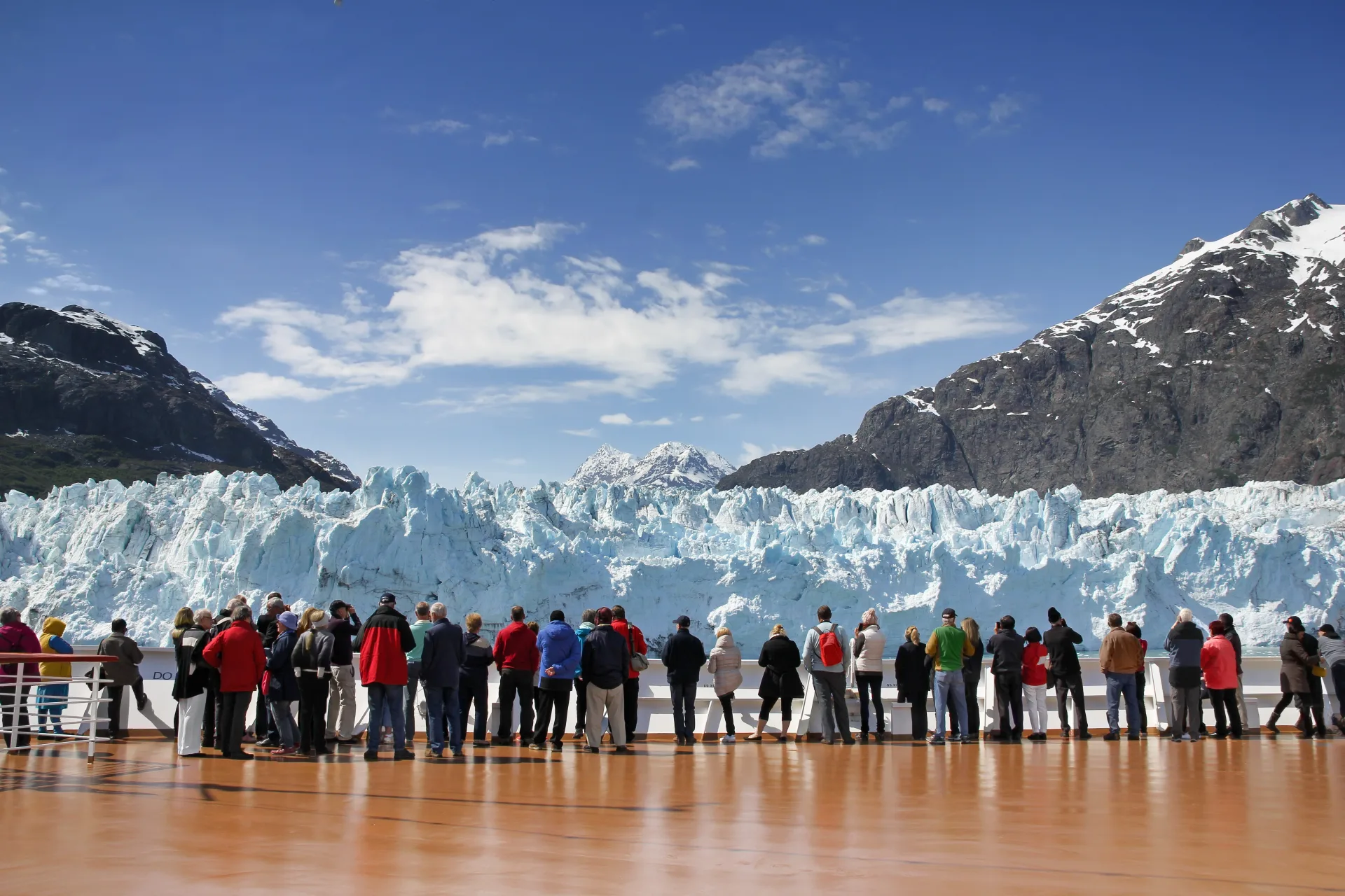 Cruise passengers on ship deck viewing a massive Glacier Bay ice wall with snow-capped mountains on an Alaska cruise.