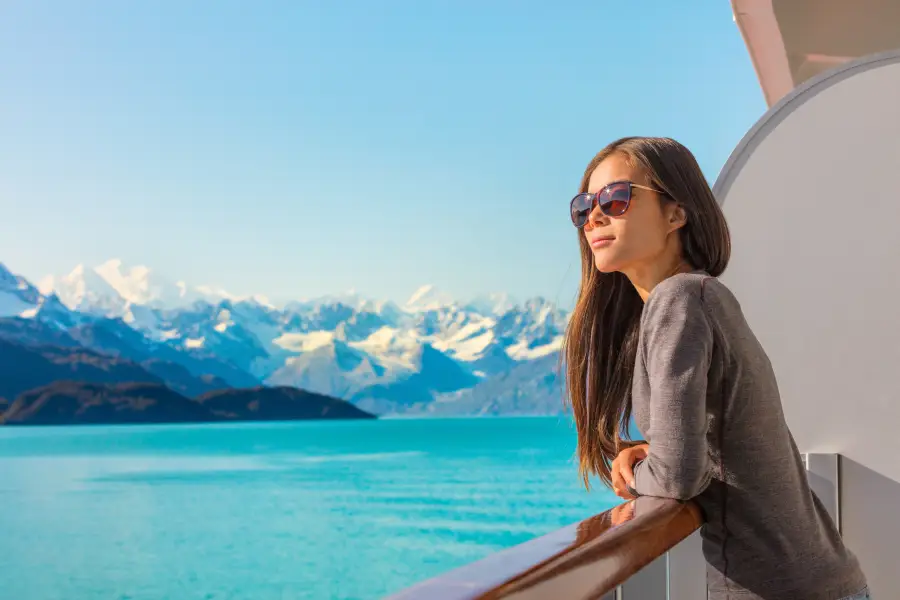Traveler on alaska cruise ship balcony gazing at snow-capped mountains over turquoise glacial waters on a clear day.