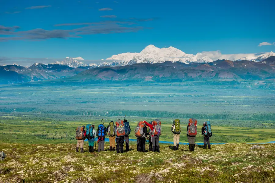 Group of backpackers standing on a tundra ridge overlooking a vast Alaska valley with Denali's snow-capped peak in the bac...