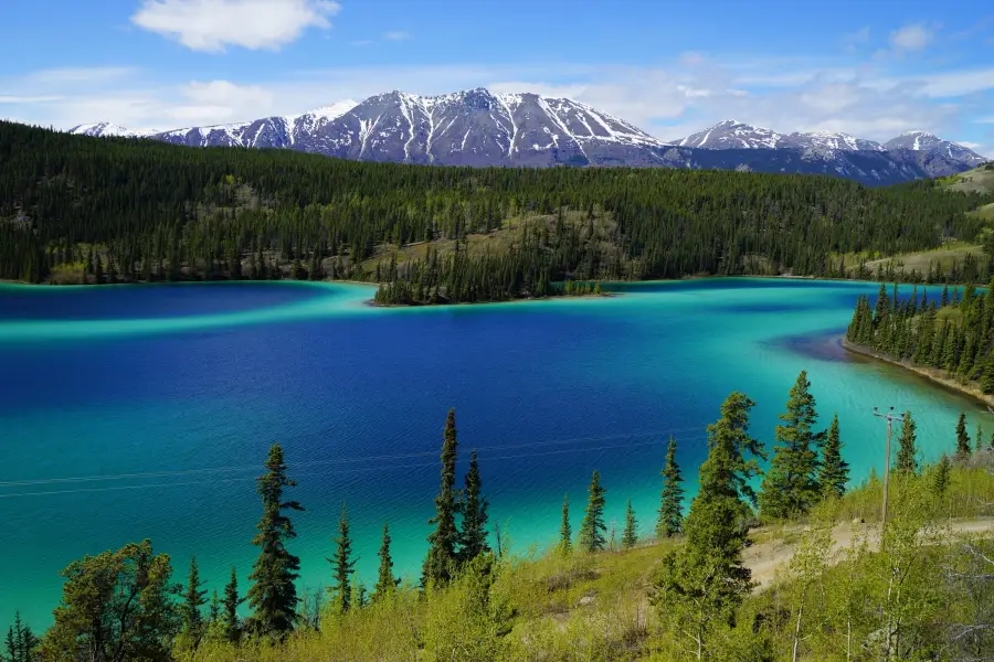 Vivid turquoise and deep blue glacial lake surrounded by boreal forest and snow-capped mountains in Alaska wilderness.