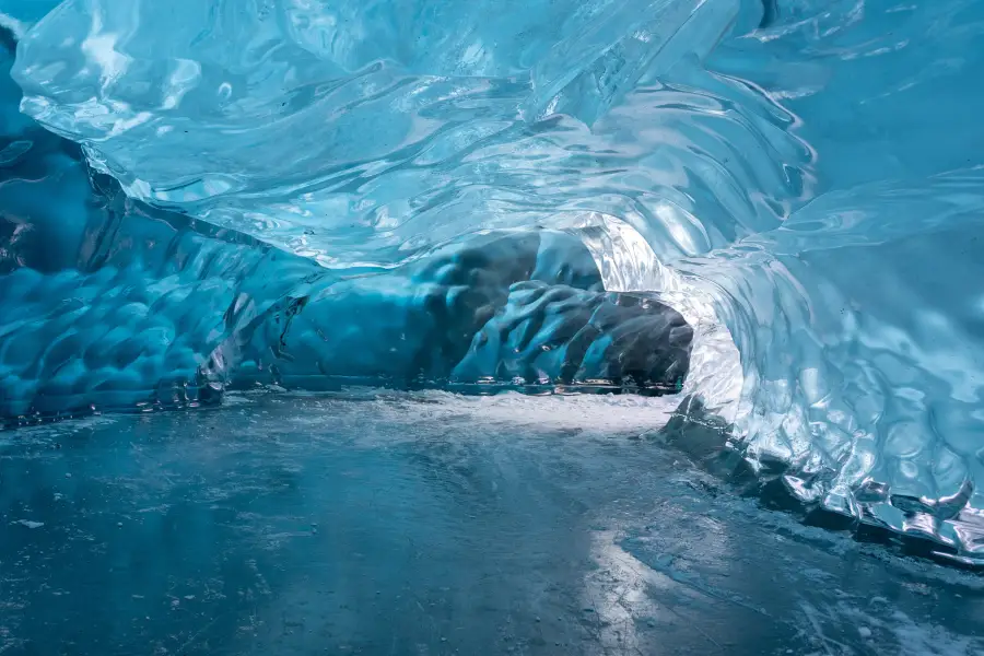Glowing blue ice cave tunnel inside an Alaska glacier; with smooth frozen walls and icy floor leading to a dark passage.