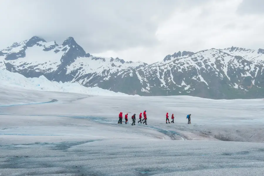 Group of hikers in red jackets trekking across an Alaska glacier with snow-capped mountains in the background.