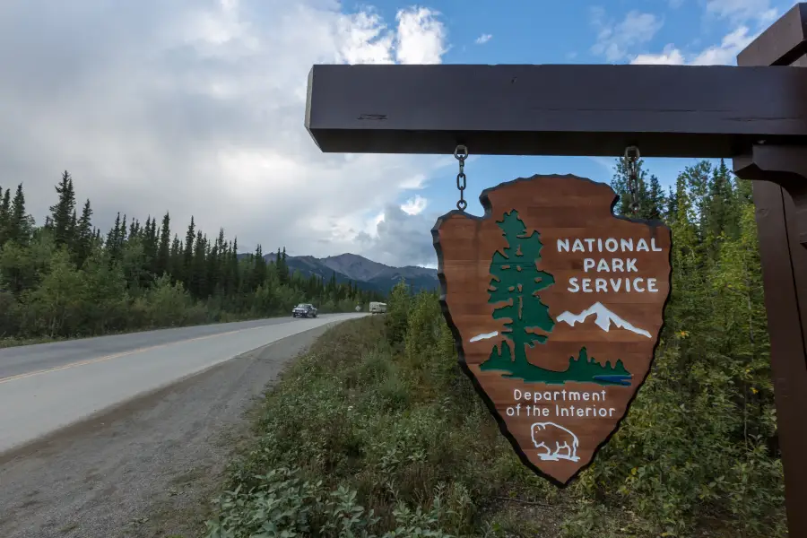 National Park Service arrowhead sign hanging roadside in Alaska; with forested highway and mountain range in background.