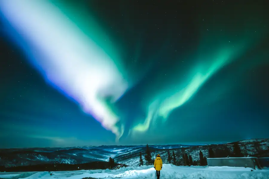 Traveler in yellow coat gazing at vivid green and white aurora borealis over snow-covered Alaska mountains at night.