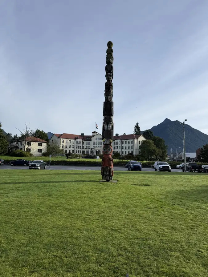 Tall carved totem pole standing on a green lawn in alaska-sitka; with a historic building and mountain in the background.