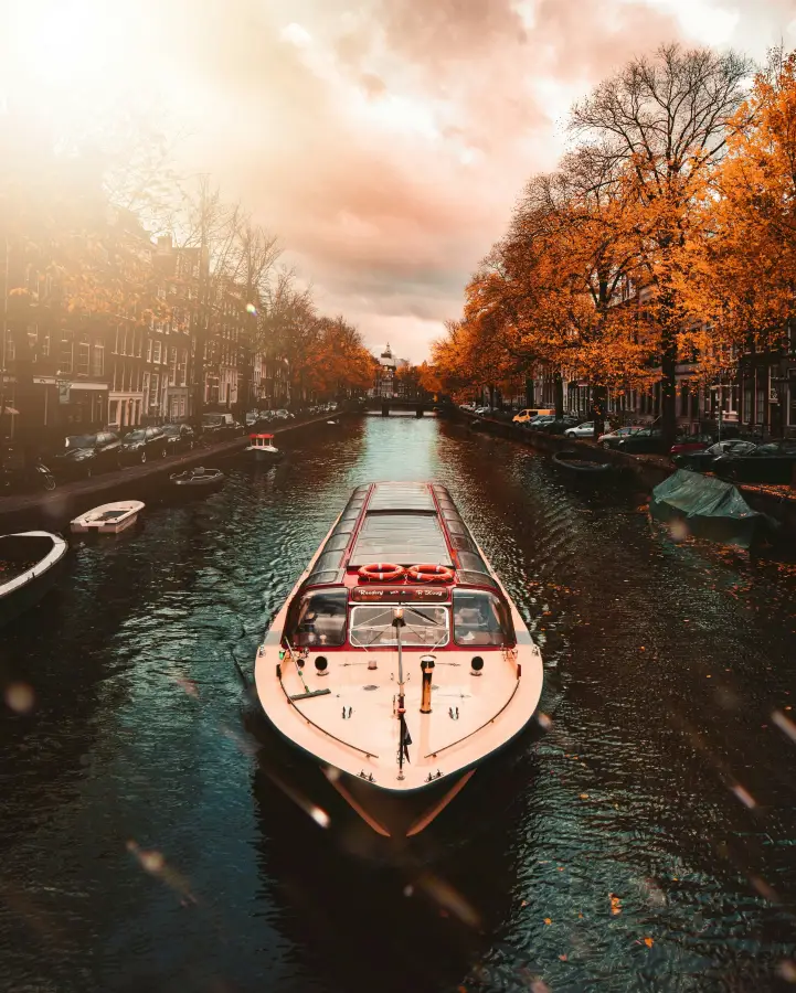 River cruise boat gliding through Amsterdam canal lined with golden autumn trees and historic canal houses at sunset.
