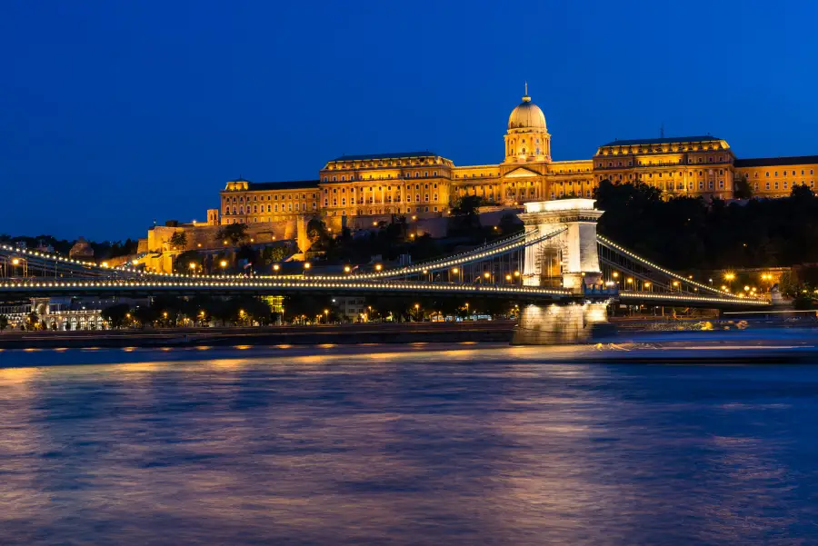Budapest river cruise view at night: illuminated Chain Bridge and Buda Castle reflecting golden light on the Danube River.