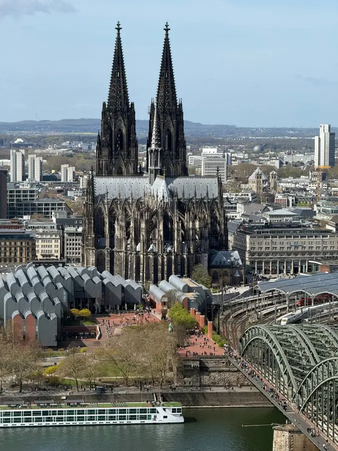 River cruise ship docked on the Rhine below Cologne Cathedral and Hohenzollern Bridge; with the city skyline beyond.