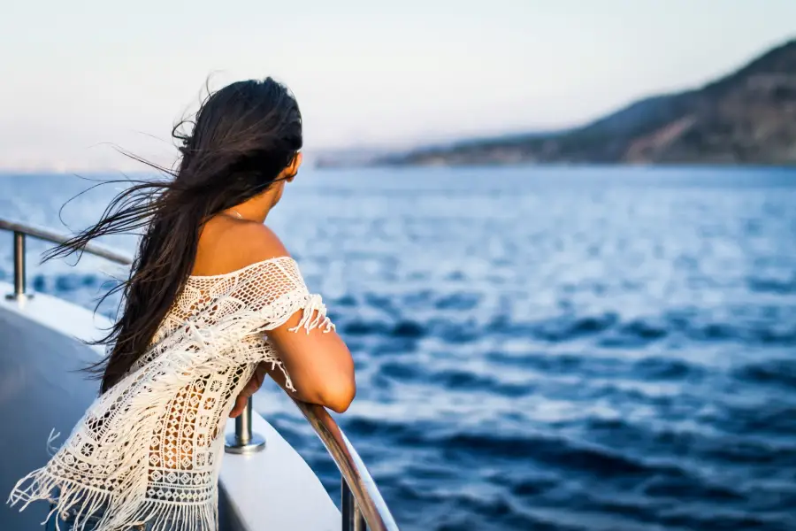 Woman in white lace top leaning on cruise ship railing; gazing at blue ocean waters with coastal hills at dusk.