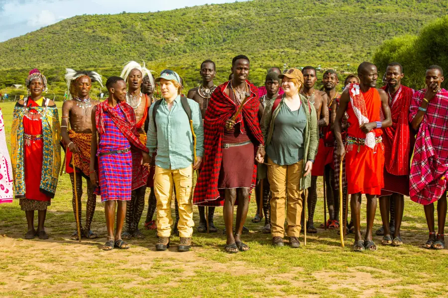 Two Western tourists stand among traditionally dressed Maasai community members during a cruise-land excursion in Kenya's...