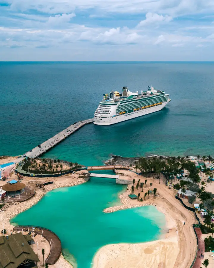 Aerial view of a Royal Caribbean cruise ship docked at a tropical island pier beside turquoise lagoon waters.