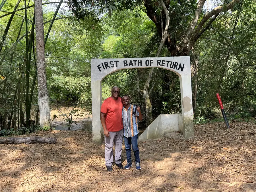 Two visitors pose under the 'First Bath of Return' arch at a heritage site during eat-mea-2024; surrounded by lush forest.