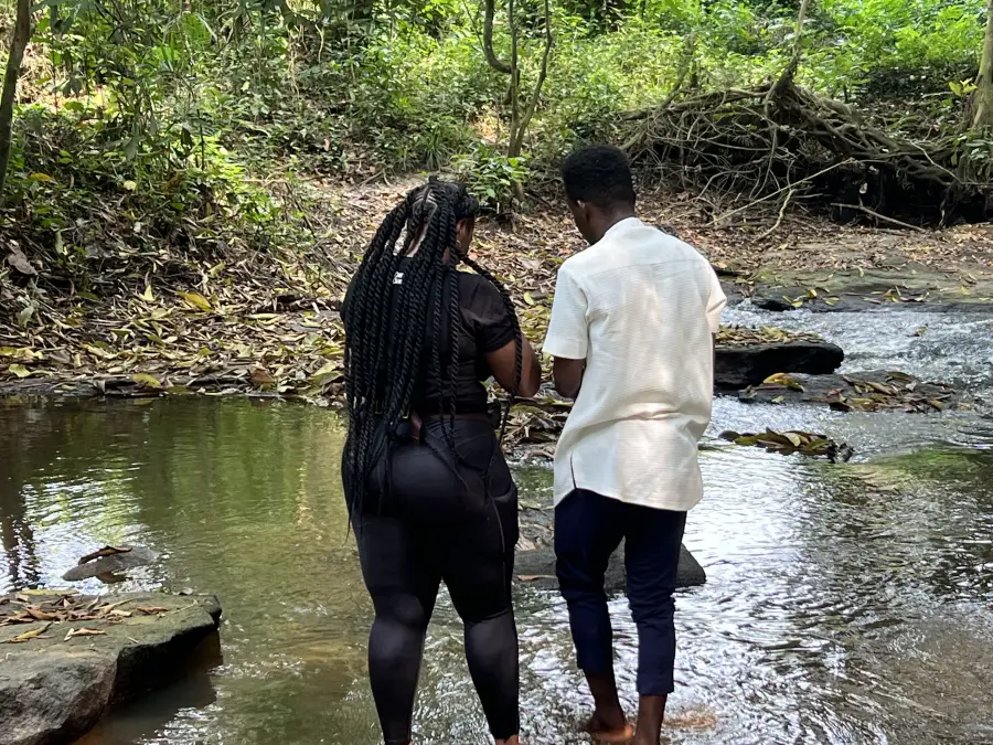 Two people stand barefoot at a forest stream during eat-mea-2024; surrounded by lush tropical greenery and rocks.