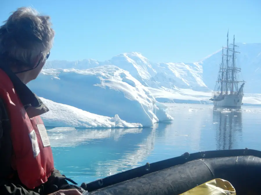 Traveler in red life vest on a Zodiac views a tall ship anchored among Antarctic icebergs during an expedition cruise.