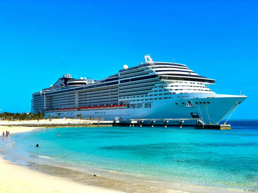 MSC Divina cruise ship docked at a tropical port with turquoise water and white sand beach under clear blue sky.