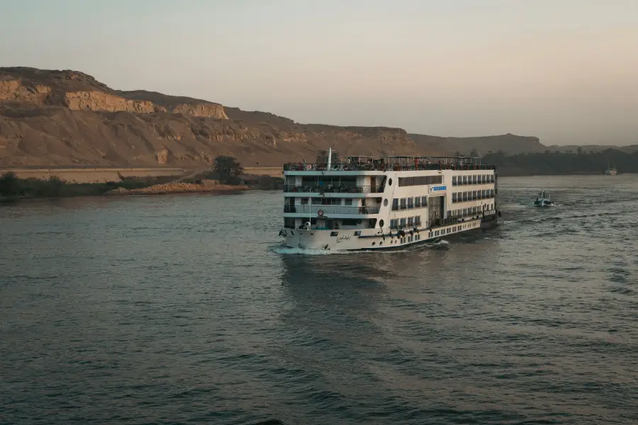 Multi-deck river cruise ship sailing the Nile at dusk with desert cliffs and palm trees in the background.