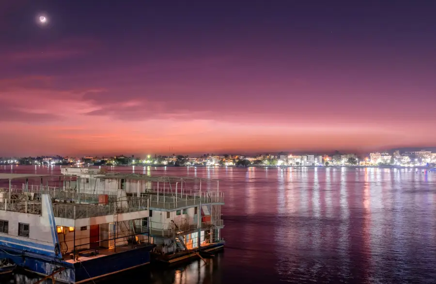 River cruise vessels docked on the Nile at twilight; city lights reflecting on calm purple-hued waters under a crescent moon.