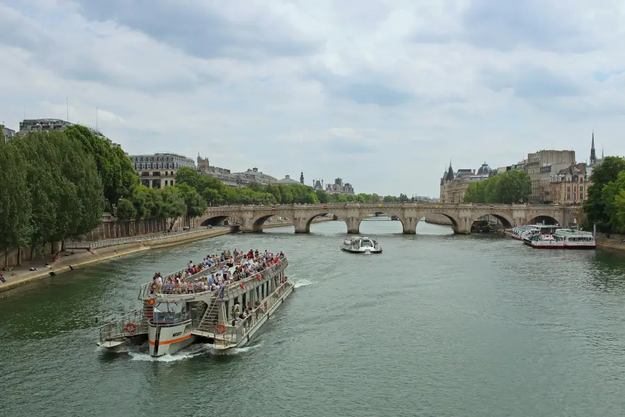 Packed river cruise boat on the Seine in Paris; with Pont Neuf arches and Haussmann buildings in the background.