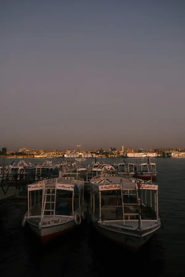 River cruise felucca boats moored on the Nile at dusk; with Luxor's illuminated skyline across the water.