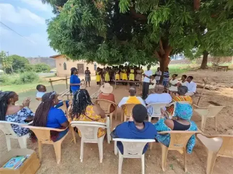 An outdoor community gathering where seated adults observe a group of school children assembled under the shade of a large, l