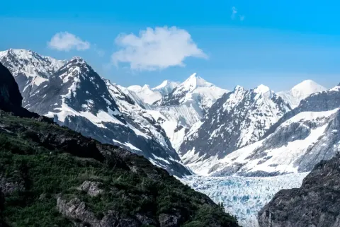 Snow-capped mountains frame a flowing glacier through a valley in Alaska under a clear blue sky.