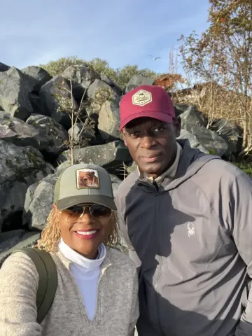 Two travelers wearing Alaska-branded caps pose near rocky shoreline in Sitka; Alaska on a sunny autumn day.