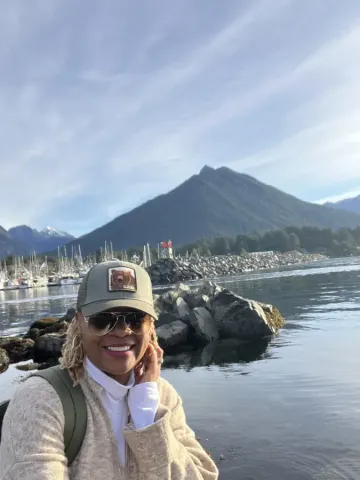 Smiling traveler in Alaska cap poses near Sitka harbor with a dramatic mountain peak and marina in the background.