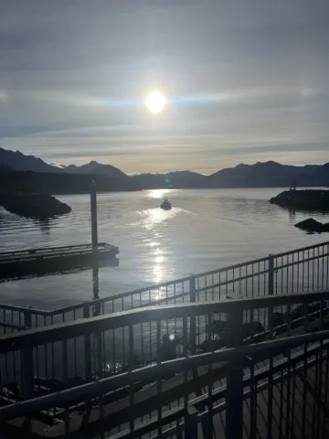 A boat glides through calm Alaska Sitka harbor waters at sunset; with mountain silhouettes and a glowing sun reflected on...