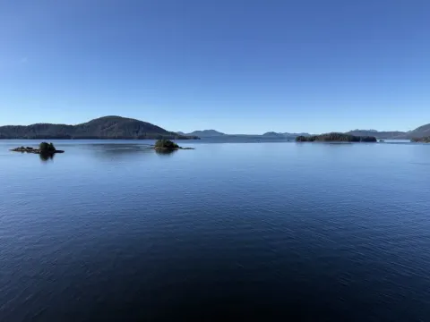 Calm blue waterway near Alaska Sitka with forested islands; rocky outcrops; and distant mountain ridges under clear skies.