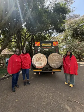 Three women in red Maasai-style capes stand smiling beside a Labusa Travel safari vehicle in Nairobi Kenya.