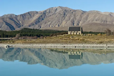 The stone Church of the Good Shepherd on the shore of Lake Tekapo, below bare mountains that reflect in the still water.