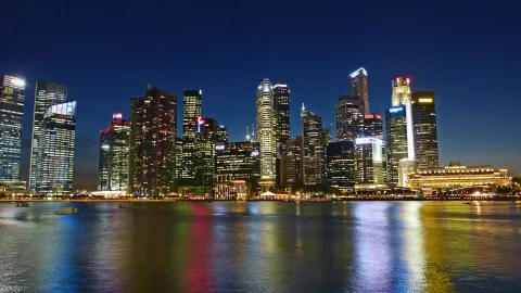 Lit towers of Singapore's Central Business District reflected in the water of Marina Bay at dusk.