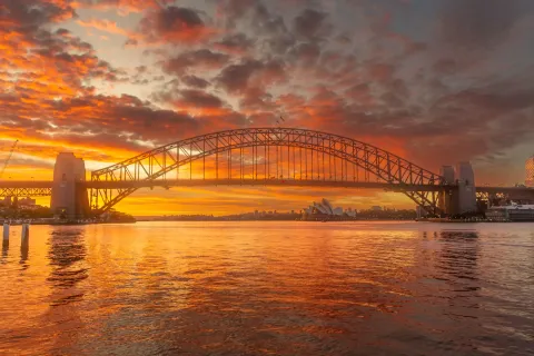 The Sydney Harbour Bridge arch at sunset, with the Opera House on the far shore beneath it.