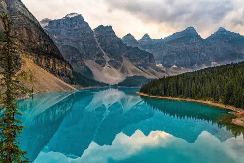 A still turquoise lake mirroring a wall of gray rock peaks, rimmed by dark spruce forest under evening cloud.
