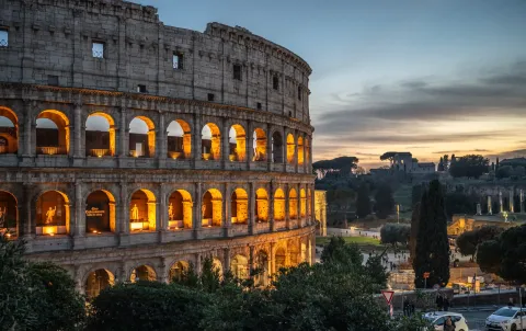 The Colosseum in Rome at dusk, its arcades lit from within, with the ruins of the Roman Forum beyond