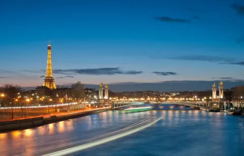 The Eiffel Tower lit at dusk above the Seine in Paris, with the gilded columns of the Pont Alexandre III to the right