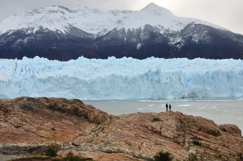 The blue ice cliff of Perito Moreno Glacier meeting the water in Los Glaciares National Park, figures on rock for scale