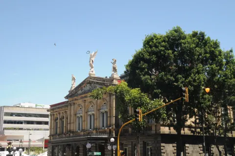 The columned stone facade and rooftop statues of the Teatro Nacional in San Jose, Costa Rica, seen past a street tree