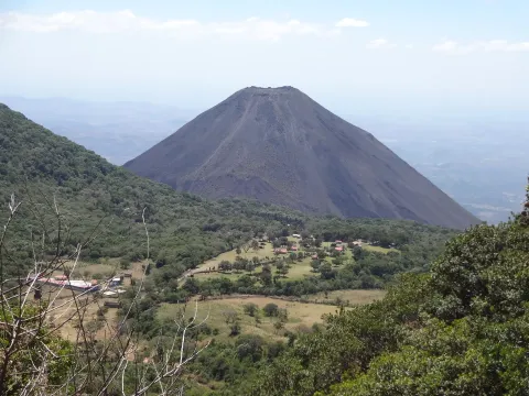 A symmetrical volcanic cone rising above forested slopes and farmland in western El Salvador under a hazy sky