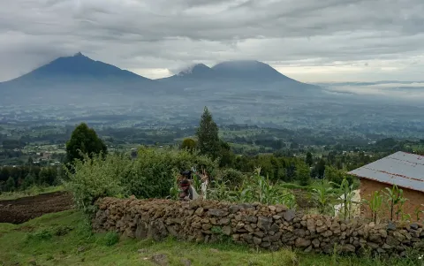 The Virunga volcanoes under cloud above cultivated fields, a lava-rock wall and a tin-roofed house on the edge of Volcanoes National Park, Rwanda.