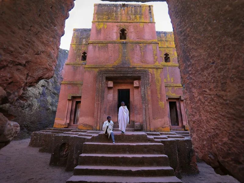 A rock-hewn church at Lalibela, Ethiopia, cut down into reddish rock so that its facade stands at the foot of an open trench, with steps leading down and a figure in white in the doorway.