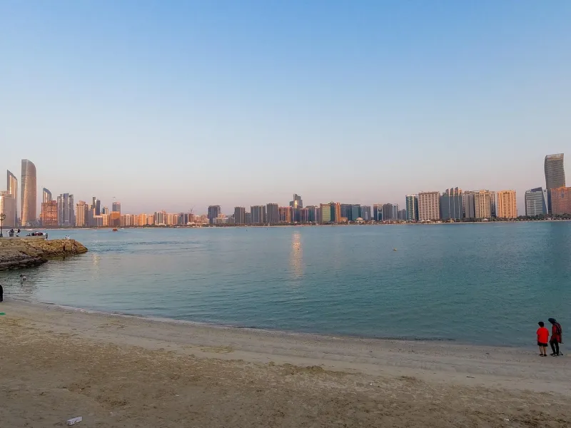 The Abu Dhabi corniche skyline seen across shallow water from a public beach at dusk