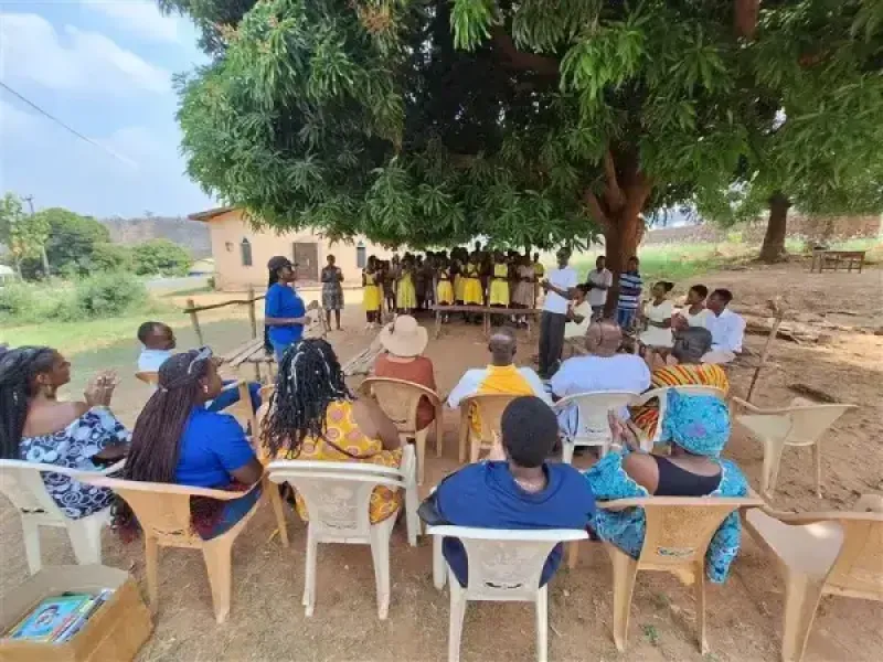 An outdoor community gathering where seated adults observe a group of school children assembled under the shade of a large, l