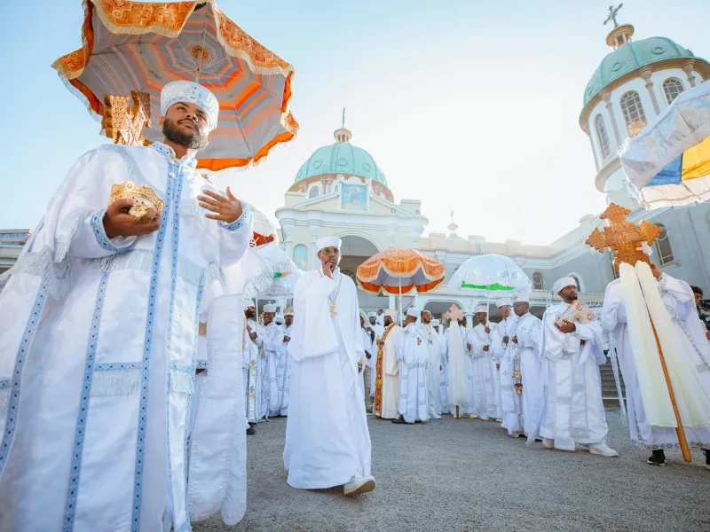 Orthodox clergy in white ceremonial robes beneath fringed ceremonial umbrellas, processing in front of a domed church.