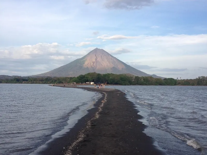 A dark volcanic sand spit running out across the water toward a symmetrical conical volcano, with a wooded shore behind it.