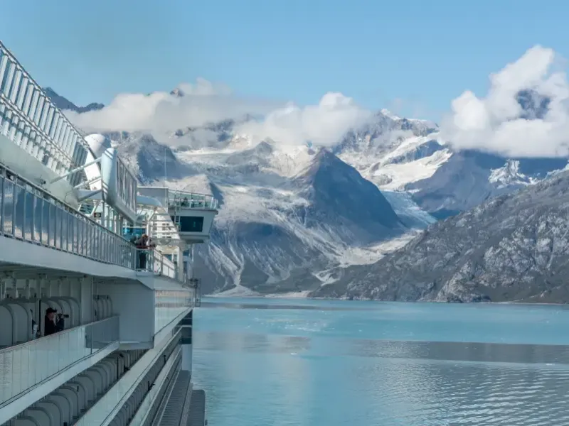 Alaska cruise ship balconies overlooking glacial Glacier Bay waters with snow-capped mountains and flowing glaciers beyond.