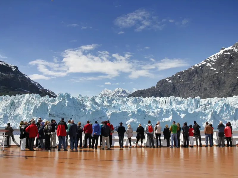 Cruise passengers on ship deck viewing a massive Glacier Bay ice wall with snow-capped mountains on an Alaska cruise.