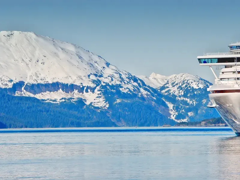 Alaska cruise ship bow navigating calm fjord waters with snow-capped forested mountains in the background.